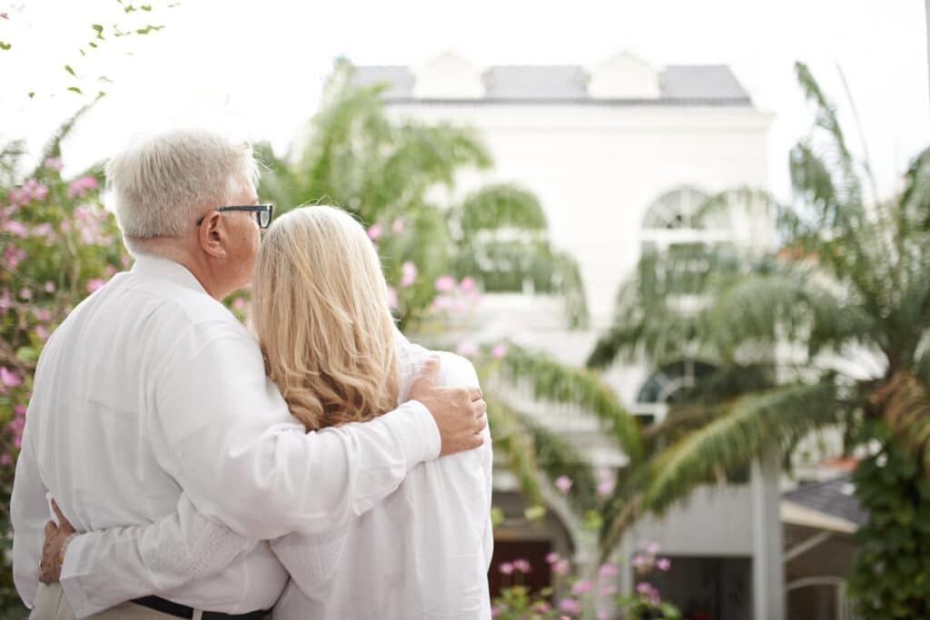 An elderly couple stands close together outdoors, lovingly embracing as they admire a large white home with palm trees and blooming flowers in the garden